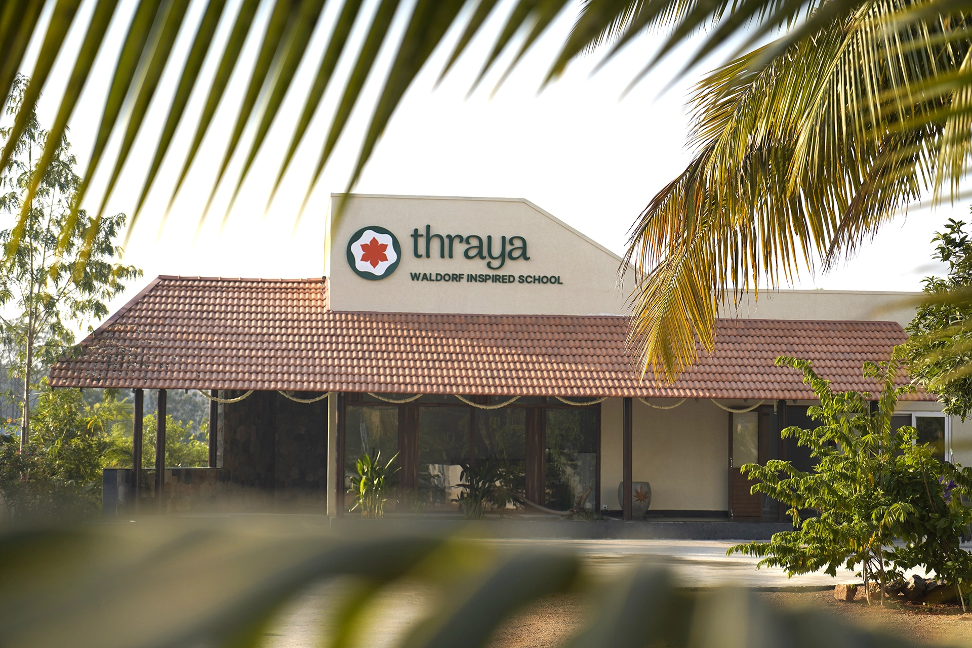 The Thraya school building in Janwada, with its tiled roof, seen through palm fronds